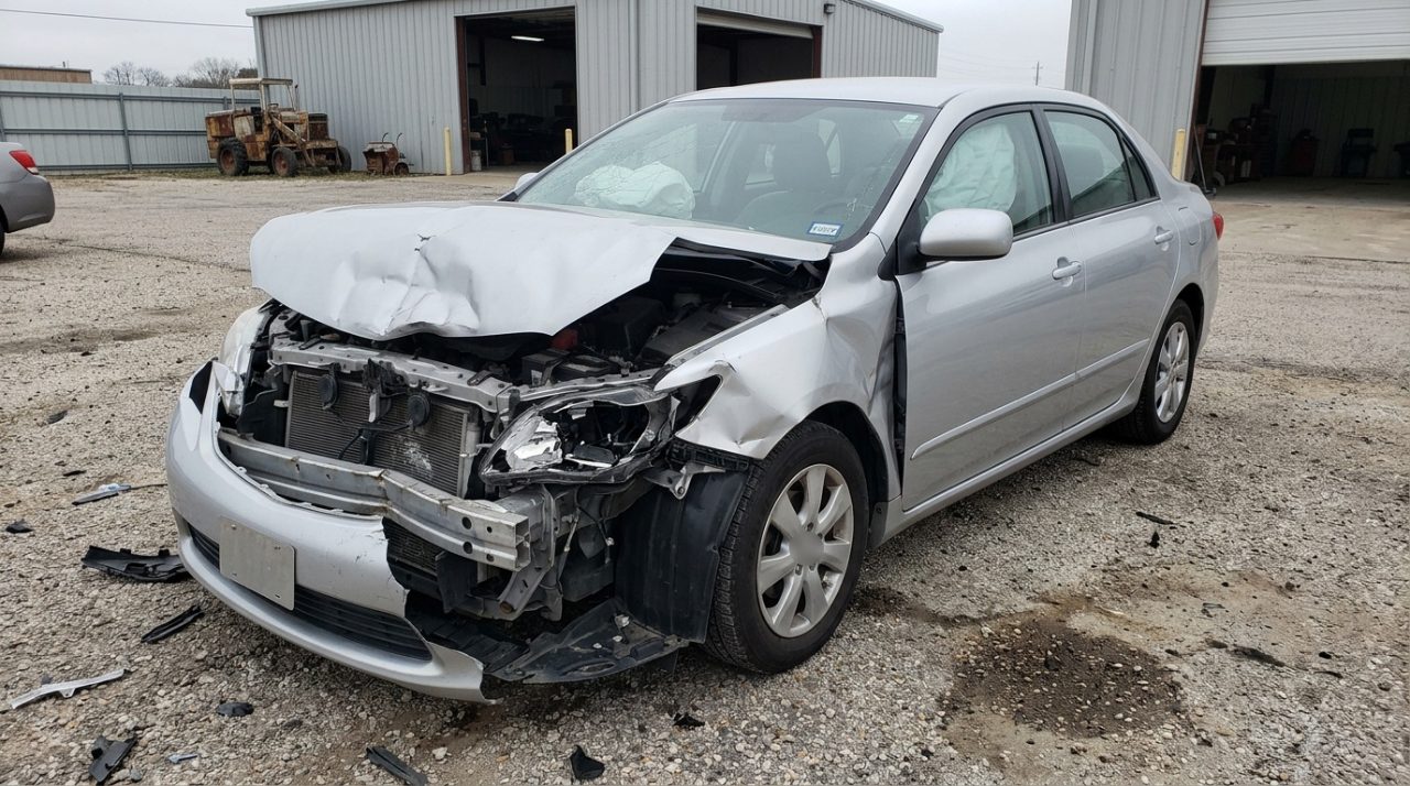 A silver sedan after a hard front-end collision: hood buckled, bumper torn away, headlight smashed and airbags deployed