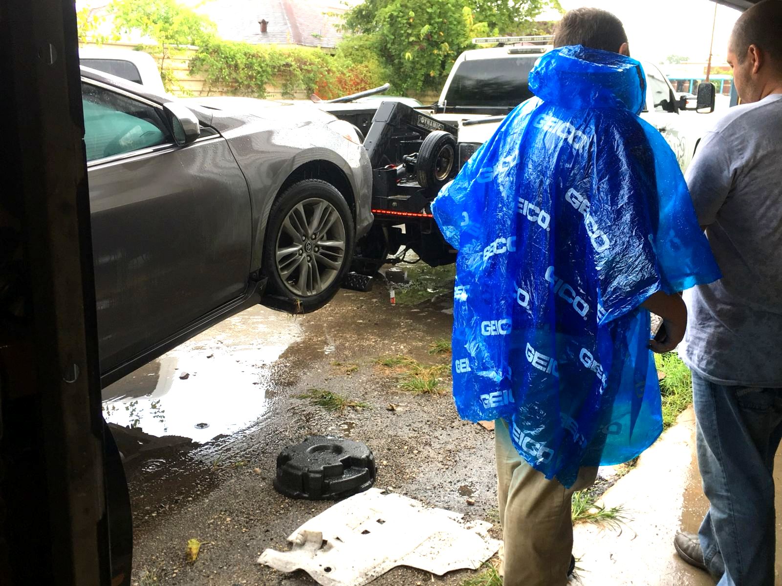 An insurance adjuster in a rain poncho watching a damaged car arrive on a tow truck at the Astro Collision shop