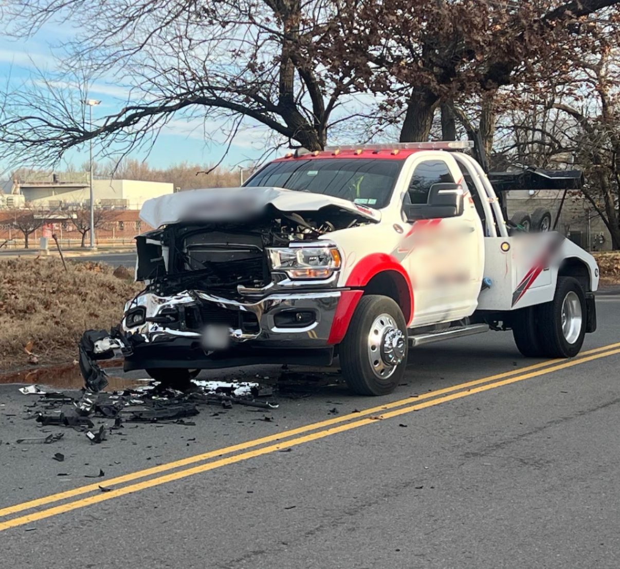 A wrecked white tow truck stopped on the road after a front-end crash, its hood crumpled and front end torn open, debris on the asphalt