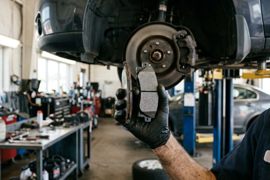 Technician holding a worn brake pad beside a new pad under a car on the lift